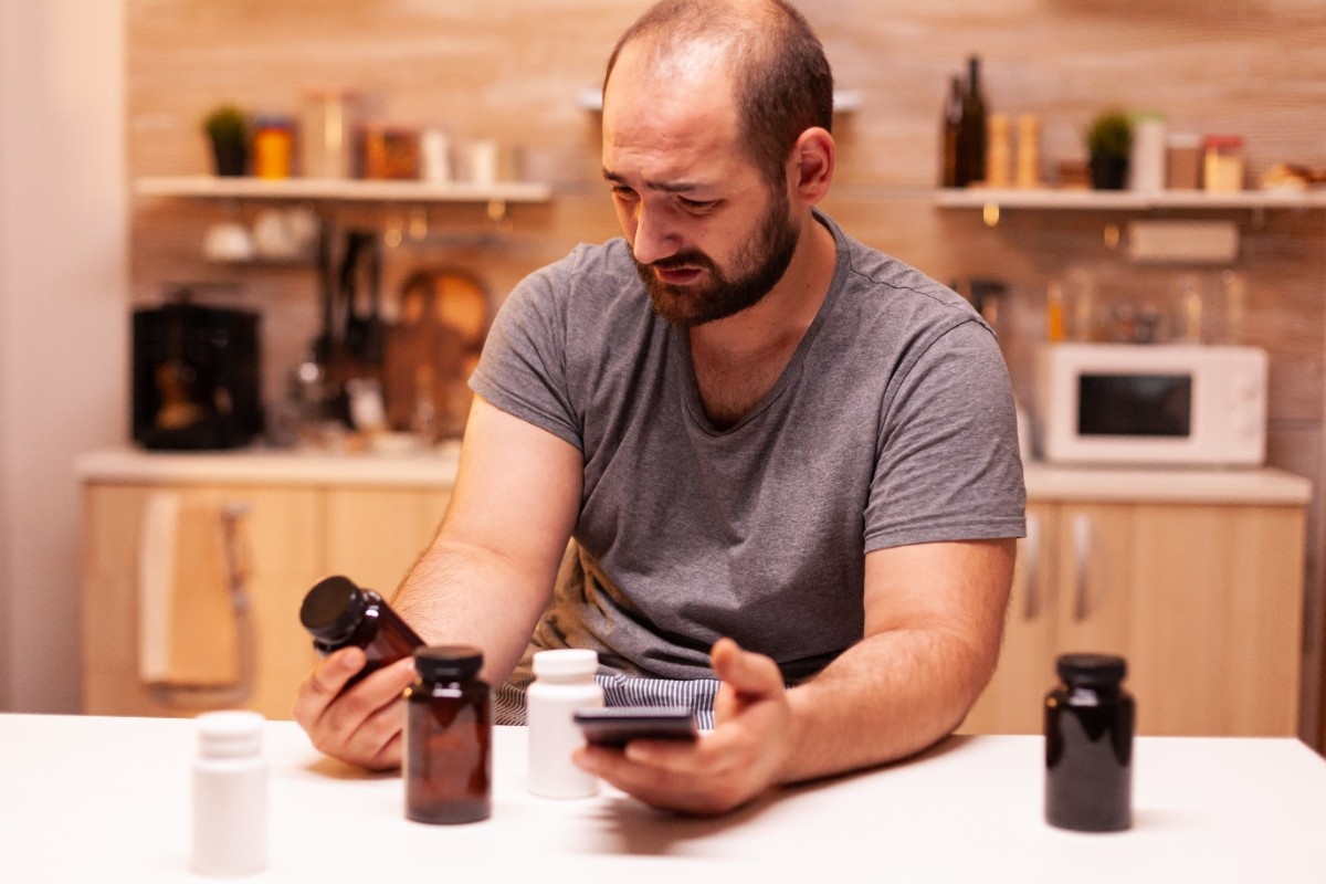 Low Testosterone in Your 30s? What You Need to Know Now 1 man with headache holding pills bottle sitting kitchen stressed tired unhappy worried person suffering migraine depression disease anxiety feeling exhausted with dizziness symptoms 1