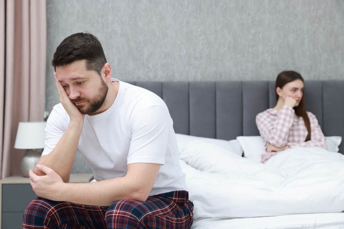 Man sitting on the edge of the bed looking distressed while his partner sits behind him, representing low testosterone signs including low libido and mood changes in men