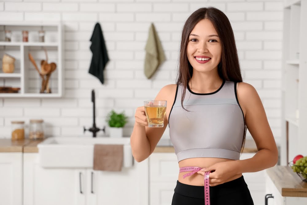 Fit woman measuring her waist while holding a healthy drink in a kitchen representing the sustained results that come from understanding how long to take weight loss medications