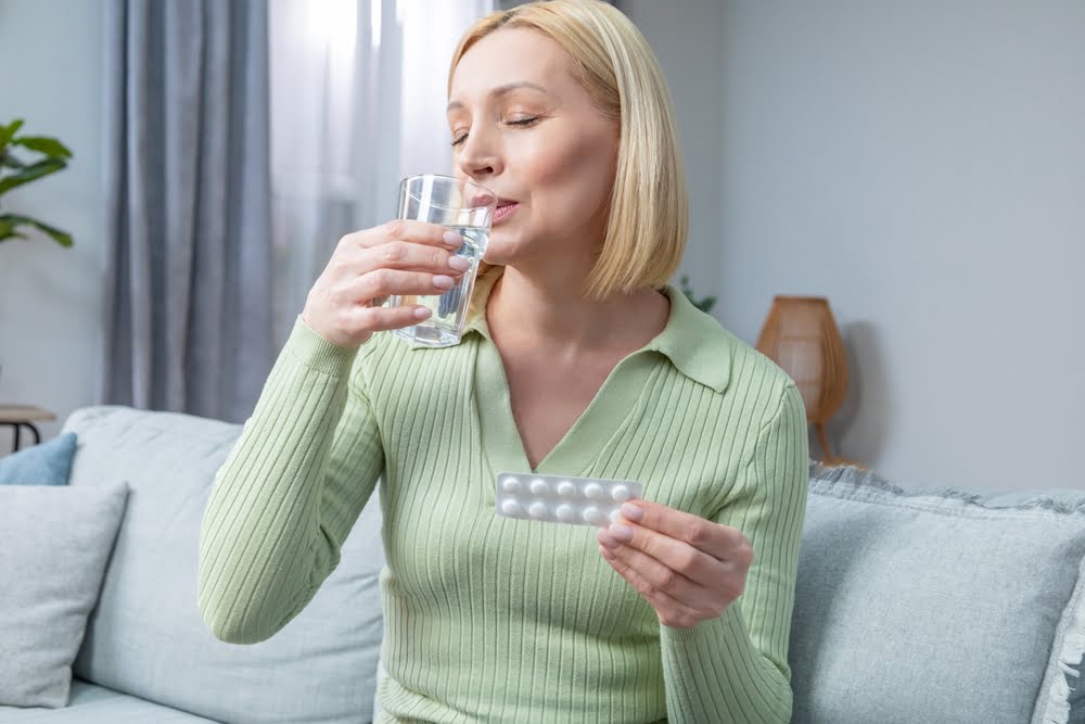 Middle-aged woman sitting on a sofa taking oral medication with water while holding a blister pack representing oral bioidentical progesterone as one of the hormones in a BHRT protocol