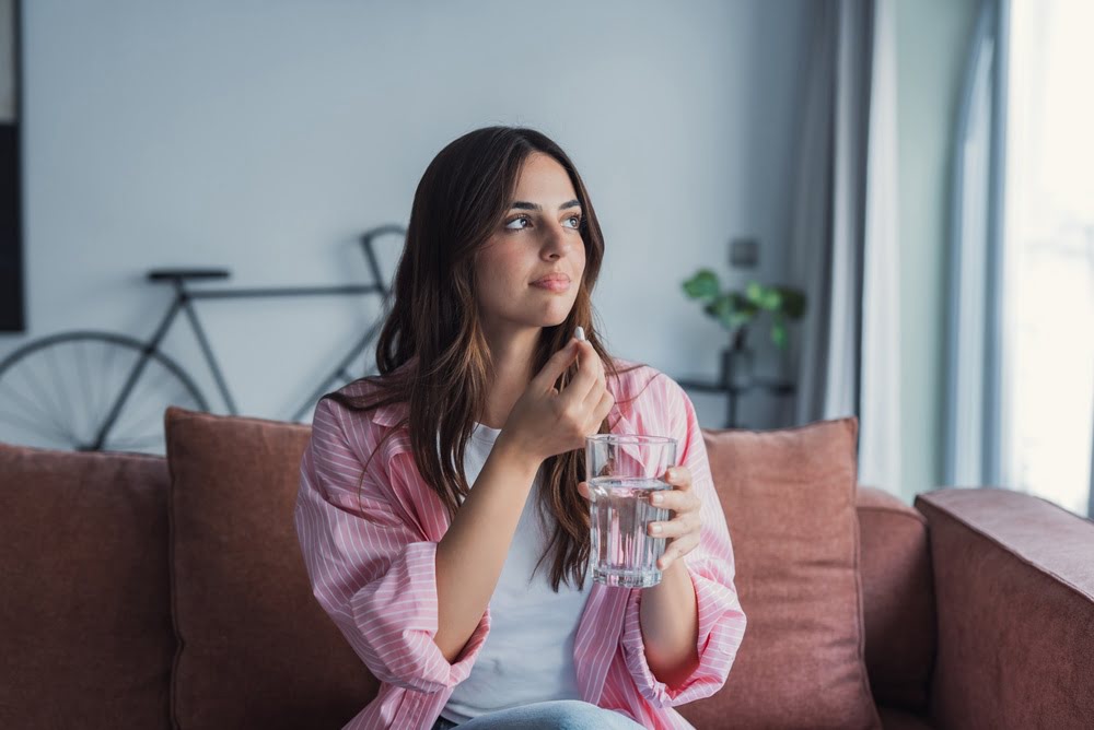 Woman sitting on a sofa holding a glass of water and an oral hormone tablet representing the long-term BHRT safety considerations involved in taking bioidentical hormone therapy consistently over time