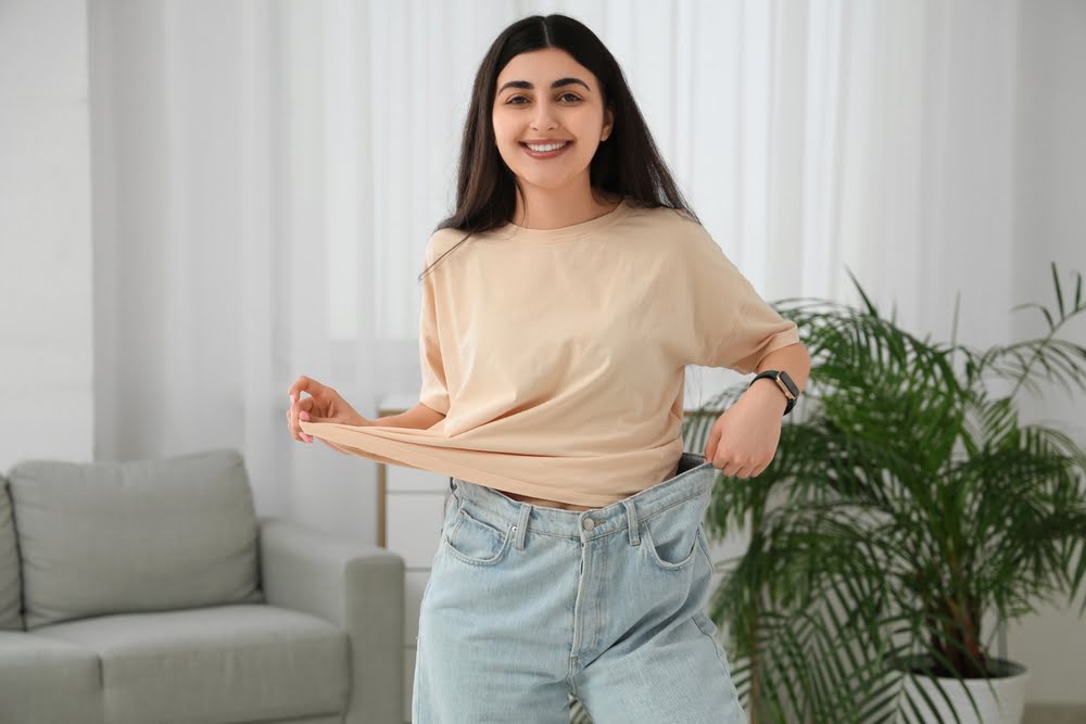 Smiling young woman pulling out the waistband of oversized jeans representing successful weight loss results achieved by a qualifying medical weight loss candidate through a supervised program