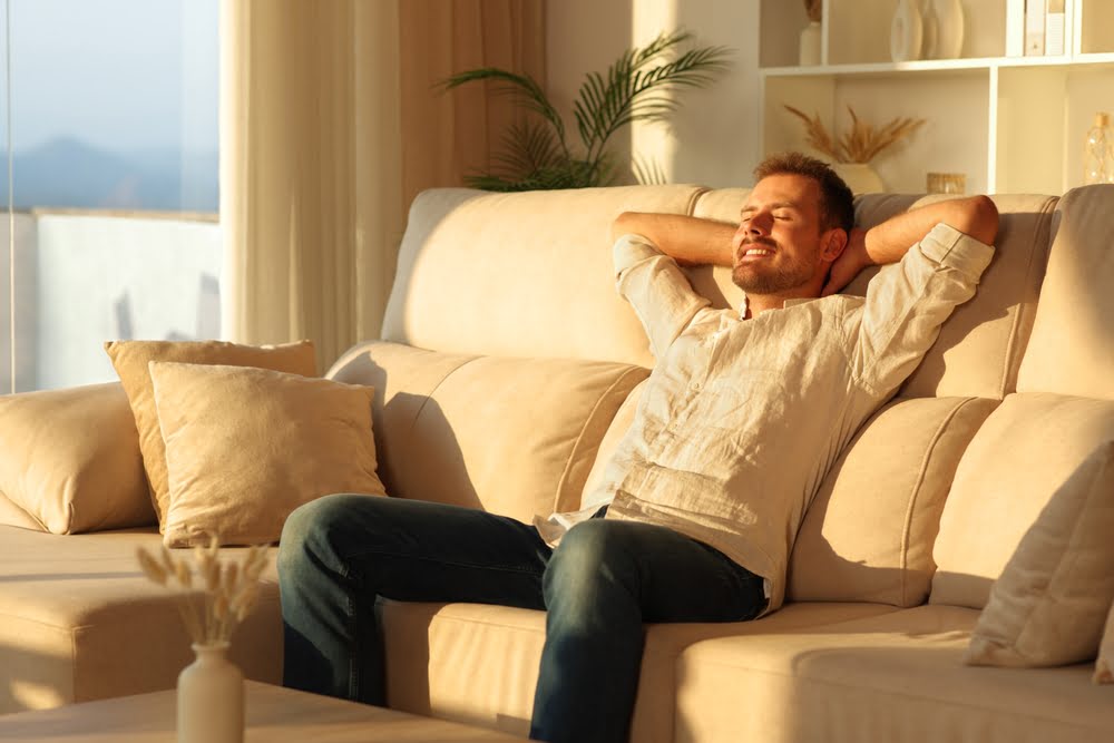 Relaxed young man resting on a sofa with eyes closed and hands behind his head in warm sunlight representing the improved emotional wellbeing and mood that TRT and mood in men can produce through testosterone optimization