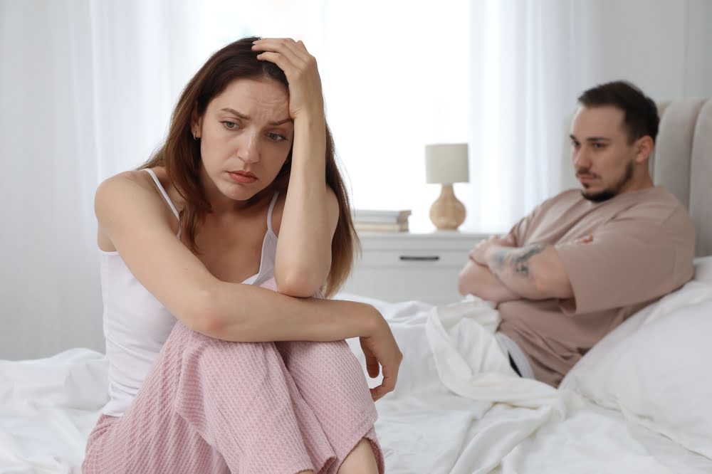 Woman sitting on the edge of a bed looking distressed while her partner sits in the background, representing the emotional and relational impact of low libido that BHRT and libido in women treatment aims to address