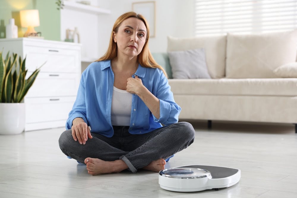 Middle-aged woman sitting on the floor looking concerned at a weighing scale representing the frustration of unexplained menopause weight gain that BHRT for menopause weight gain is designed to address by correcting the hormonal drivers behind it